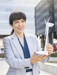 Female professional smiling while holding wind turbine model in office park - JCCMF02036