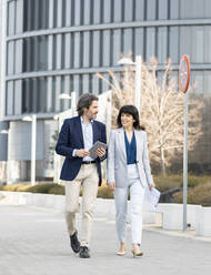 Male and female professionals discussing while walking on footpath - JCCMF02055