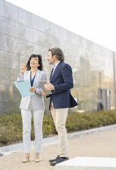 Businesswoman with clipboard gesturing while standing by male colleague on footpath - JCCMF02058