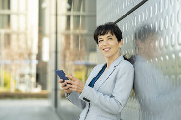 Smiling entrepreneur with mobile phone day dreaming while leaning on metal wall - JCCMF02081