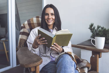 Lächelnde Frau hält ein Buch und sitzt auf einem Stuhl auf dem Balkon - OIPF00555
