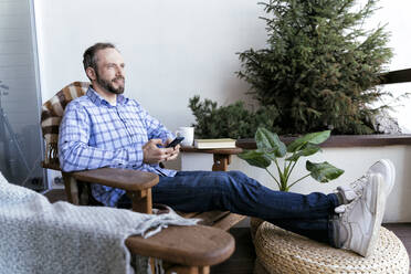 Thoughtful man looking away holding smart phone while sitting on chair on balcony - OIPF00556