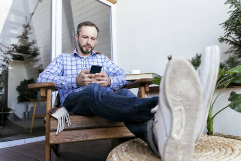 Gut aussehender Mann benutzt sein Smartphone, während er auf einem Stuhl auf dem Balkon sitzt - OIPF00557