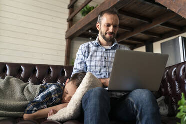 Mann benutzt Laptop, während sein Sohn zu Hause auf dem Sofa schläft - OIPF00571
