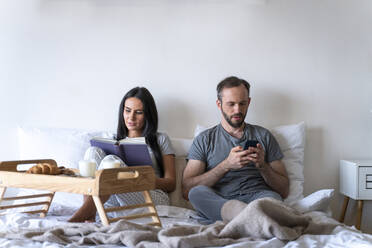 Woman reading book sitting with man using smart phone while having breakfast on bed at home - OIPF00594