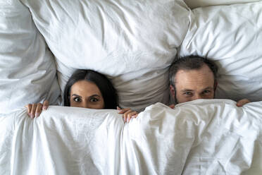 Couple hiding faces with blanket while lying on bed at home - OIPF00598