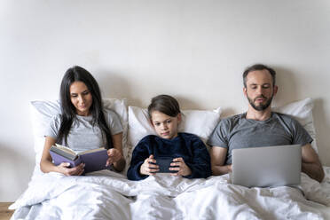 Woman reading book while father and son using wireless technologies in bed at home - OIPF00601