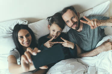 Father and son gesturing while woman taking selfie lying on bed at home - OIPF00603