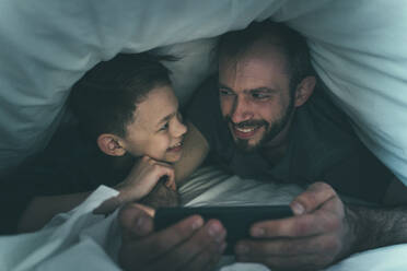Vater und Sohn schauen sich an, während sie zu Hause unter der Bettdecke ihr Smartphone benutzen - OIPF00612