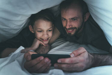 Son and father looking at smart phone under blanket at home - OIPF00613