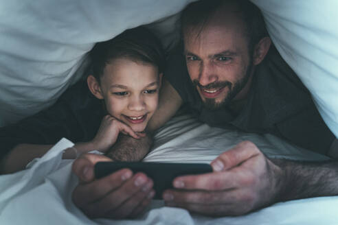 Sohn und Vater schauen zu Hause unter der Bettdecke auf ihr Smartphone - OIPF00613