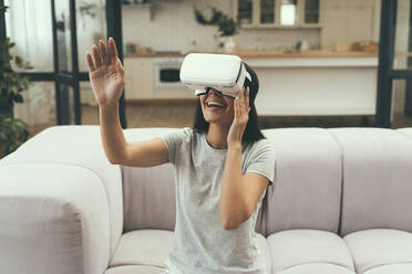 Lächelnde Frau, die mit einem Virtual-Reality-Headset gestikuliert, während sie zu Hause auf dem Sofa sitzt - OIPF00621