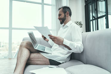 Geschäftsmann mit Laptop, der ein Dokument hält, während er zu Hause auf dem Sofa sitzt - OIPF00626