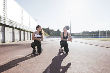 Smiling female friends crouching on training grounds during sunny day - RSGF00644