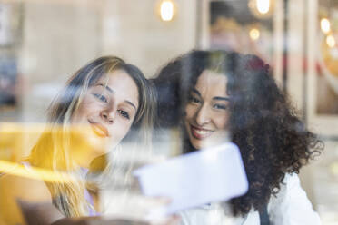Glückliche Freundinnen, die ein Selfie mit ihrem Smartphone machen, gesehen durch ein Glas im Restaurant - PNAF01498