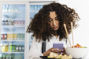 Junge Frau mit lockigem Haar, die im Restaurant sitzend eine Textnachricht über ihr Smartphone verschickt - PNAF01532