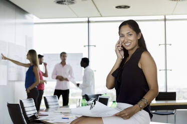 Female professional talking on mobile phone while sitting on table with male and female colleagues in background at office - BMOF00564