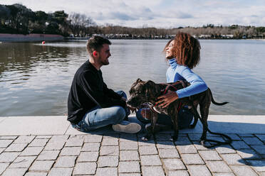 Smiling girlfriend with boyfriend sitting by dog on footpath by lake - EBBF03428