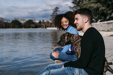 Lächelndes junges Paar sitzt mit Hund am See an einem sonnigen Tag - EBBF03429
