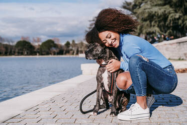 Woman embracing pet while crouching on footpath by lake - EBBF03431