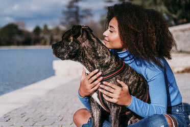 Frau mit lockigem Haar schaut weg, während sie mit einem American Pit Bull Terrier an einem sonnigen Tag sitzt - EBBF03433