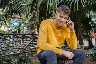 Smiling man talking on mobile phone while sitting on bench at park - DLTSF01822