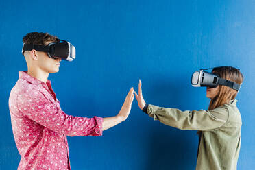 Friends with virtual reality headset stretching hands towards each other by blue wall - DLTSF01842