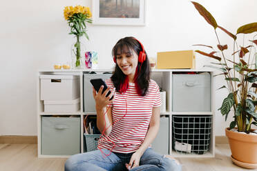 Smiling woman with headphones using smart phone while sitting at home - TCEF01810