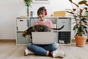 Junge Frau mit Kopfhörern sitzt mit Laptop und Katze zu Hause - TCEF01811