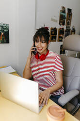 Young woman with headphones and laptop talking on mobile phone at home - TCEF01817