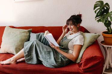 Smiling woman with eyeglasses reading book while sitting on sofa at home - TCEF01822