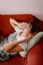 Young woman listening music while lying on red sofa - TCEF01824