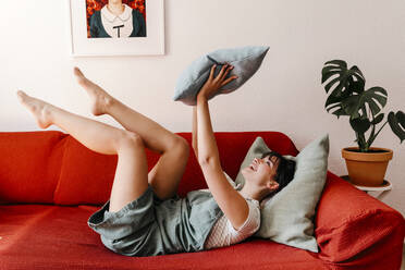 Glückliche Frau spielt mit Kissen, während sie auf dem Sofa liegt - TCEF01826