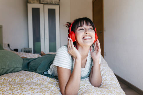 Beautiful woman wearing headphones smiling while lying on front in bed - TCEF01833