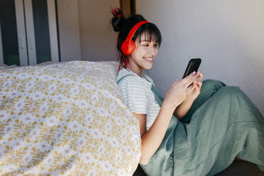 Junge Frau mit Kopfhörern, die ein Mobiltelefon benutzt, während sie im Schlafzimmer sitzt - TCEF01834