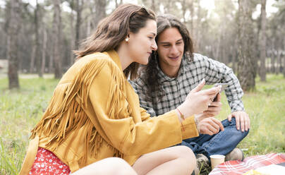 Woman showing digital tablet to young man while sitting at forest - JCCMF02119
