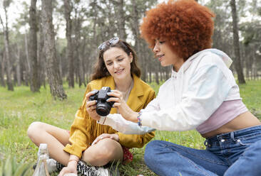 Female friends looking at photograph in camera while sitting at forest - JCCMF02124