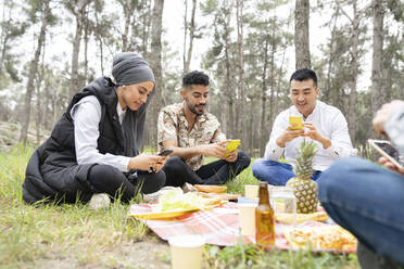 Männliche und weibliche Freunde benutzen ein Smartphone, während sie im Wald sitzen - JCCMF02130