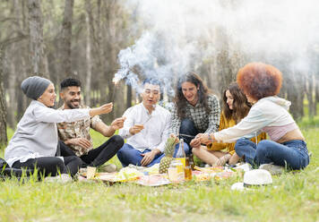 Playful male and female friends holding sparkler while sitting at forest - JCCMF02158