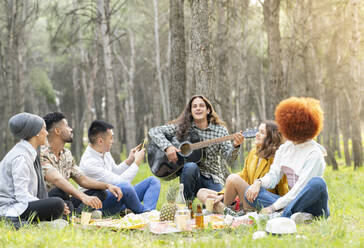 Mann mit männlichen und weiblichen Freunden spielt Gitarre beim Picknick im Wald - JCCMF02160