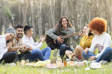 Mann mit männlichen und weiblichen Freunden spielt Gitarre beim Picknick im Wald - JCCMF02161