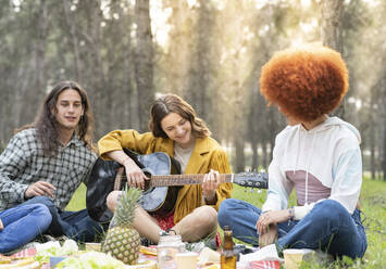 Frau mit männlichem und weiblichem Freund spielt Gitarre beim Picknick im Wald - JCCMF02164