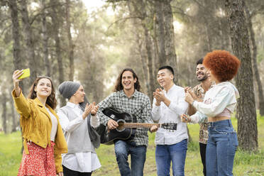 Happy woman taking selfie on mobile phone with male and female friends in forest - JCCMF02178