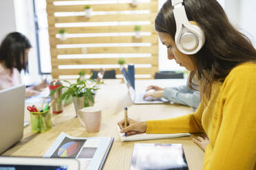 Berufstätige Frau mit Kopfhörern beim Schreiben im Büro - JSMF02149