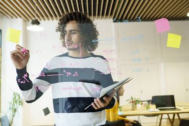 Curly haired creative businessman holding clipboard while writing on glass wall in office - JSMF02165