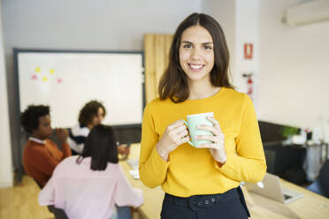 Lächelnde kreative weibliche Fachkraft, die eine Tasse im Büro hält - JSMF02190