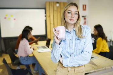 Blond hair creative businesswoman holding coffee mug while leaning at office desk - JSMF02194