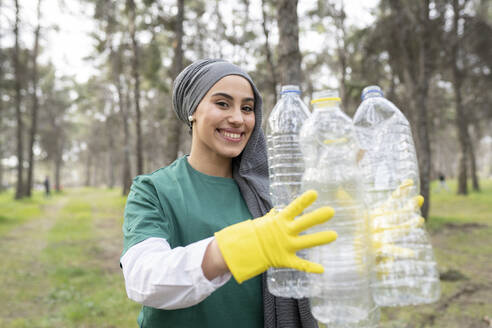 Lächelnde weibliche Freiwillige mit Schutzhandschuh, die eine Plastikflasche im Wald hält - JCCMF02198