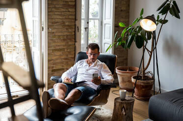 Businessman sitting in armchair and taking notes in organizer while browsing smartphone and checking messages - ADSF23627