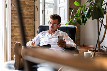 Businessman sitting in armchair and taking notes in organizer while browsing smartphone and checking messages - ADSF23629